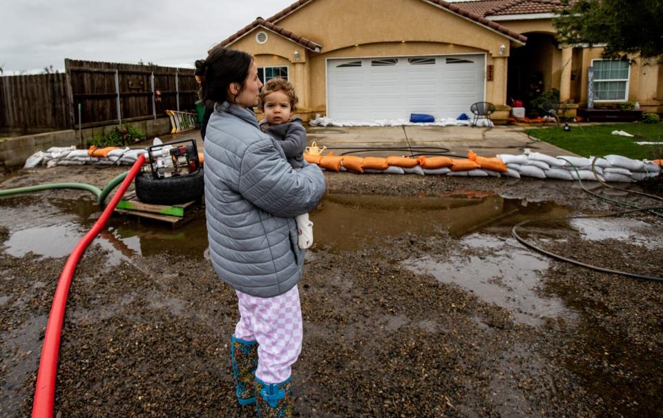 A woman holds a young boy in a mud-covered street next to pumps, hoses and sandbags in front of a house