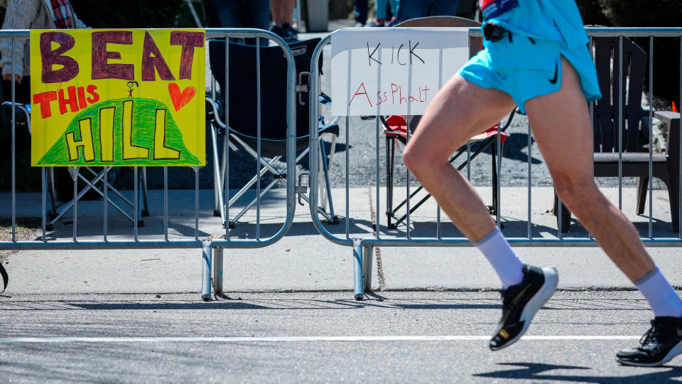 A runner in the Boston Marathon runs past spectator signs hung on metal fence. The signs read “Beat this hill” and “Kick AssPhalt”