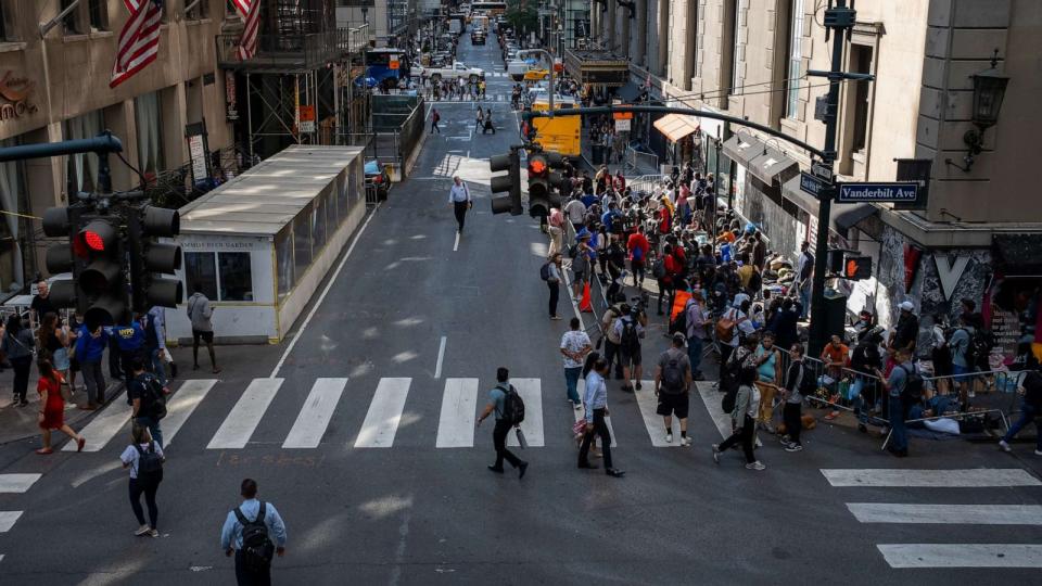 PHOTO: People walk past migrants gathered outside of the Roosevelt Hotel where dozens of recently arrived migrants have been camping out as they try to secure temporary housing, Aug. 2, 2023, in New York. (Alexi Rosenfeld/Getty Images)