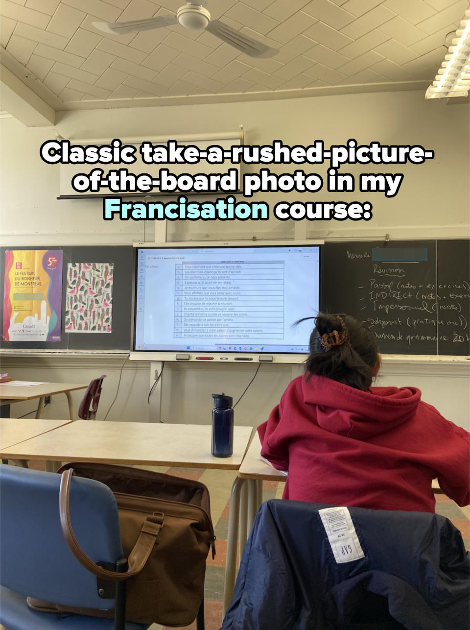 Person in a red hoodie and black hair bun sits in a classroom facing a whiteboard with a projected presentation