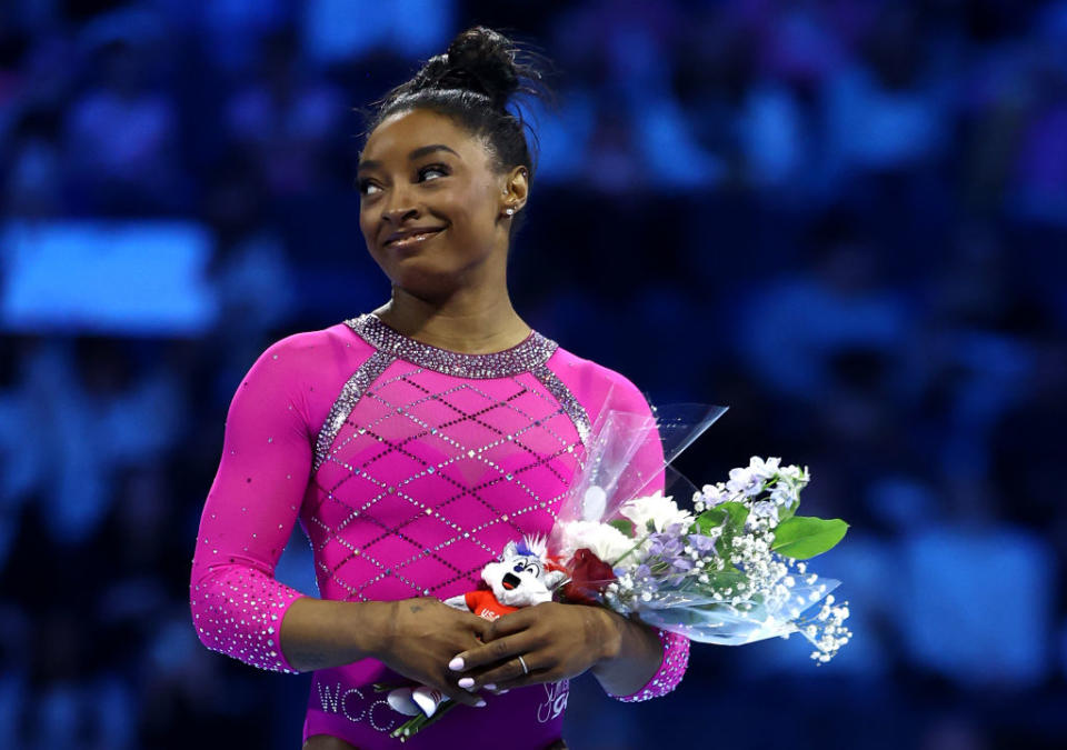 Simone Biles, in a bejeweled leotard, holds a bouquet of flowers and a small plush toy at a gymnastics event