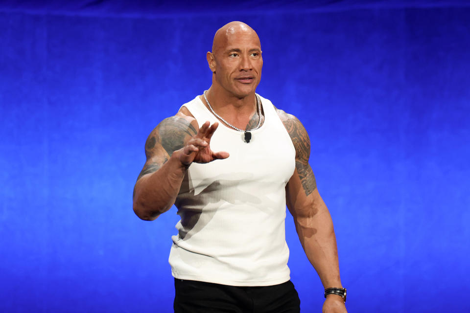 Dwayne Johnson is speaking on stage wearing a sleeveless white shirt and black pants. He has visible tattoos and is gesturing with one hand