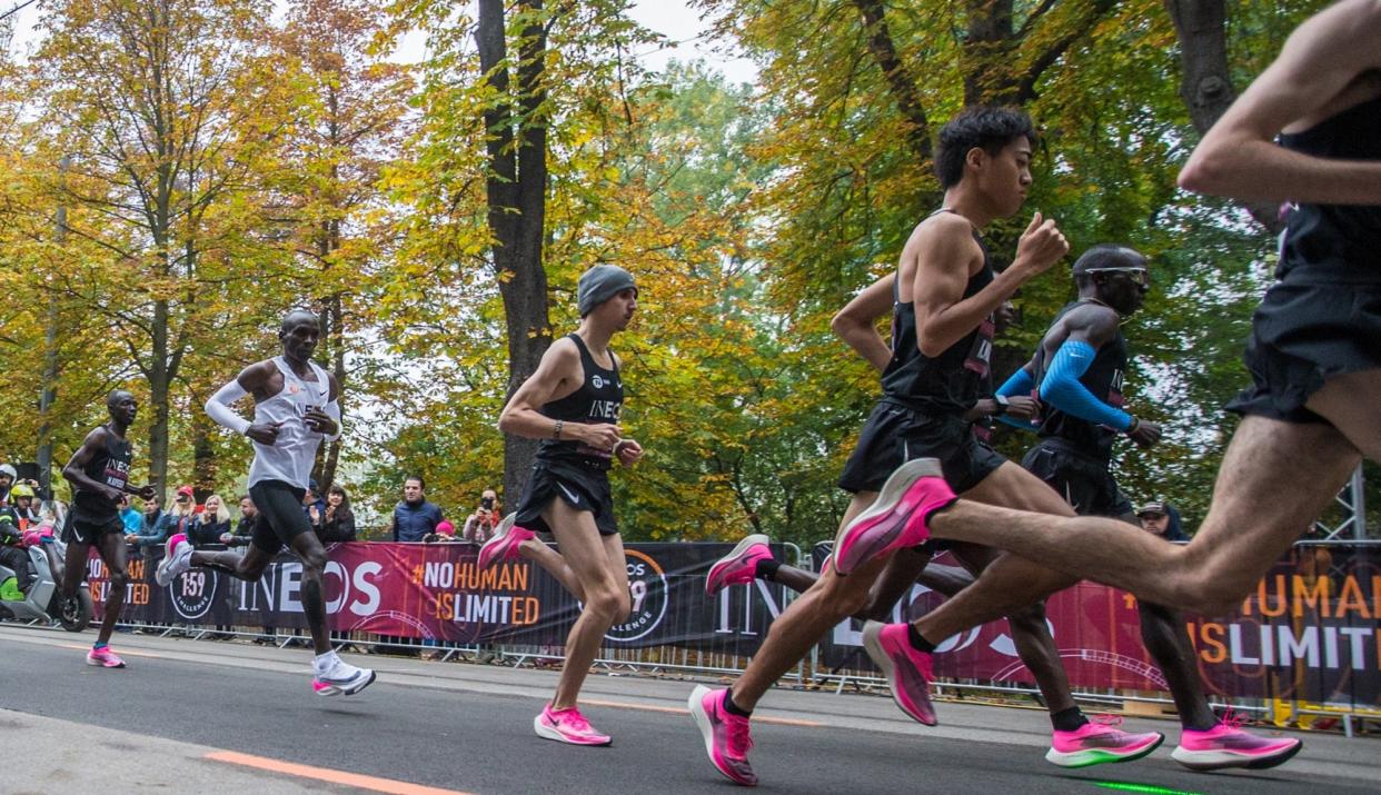 Runners forming a V-shape with marathoner Eluid Kipchoge in the rear, on a road beside trees.