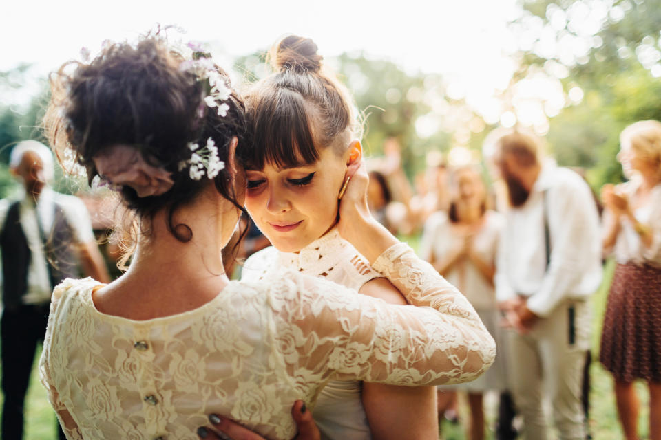 A bride and her partner share an intimate embrace during their wedding ceremony in an outdoor setting, surrounded by friends and family in the background