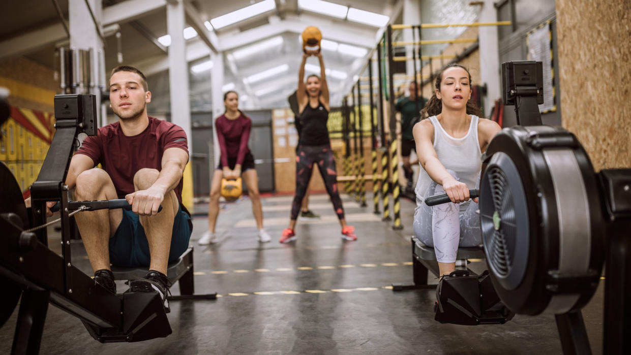  Two people rowing on rowing machines in the foreground, two people working out with kettlebells in the background 