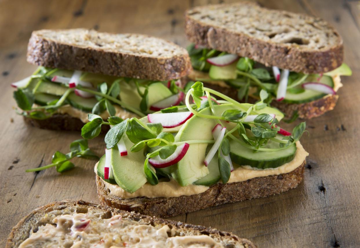 Three healthy vegetarian sandwich's on a rustic wood background. Sandwich's are on multigrain bread with roasted red pepper hummus , radish's ,cucumber , pea shoots and avocado.