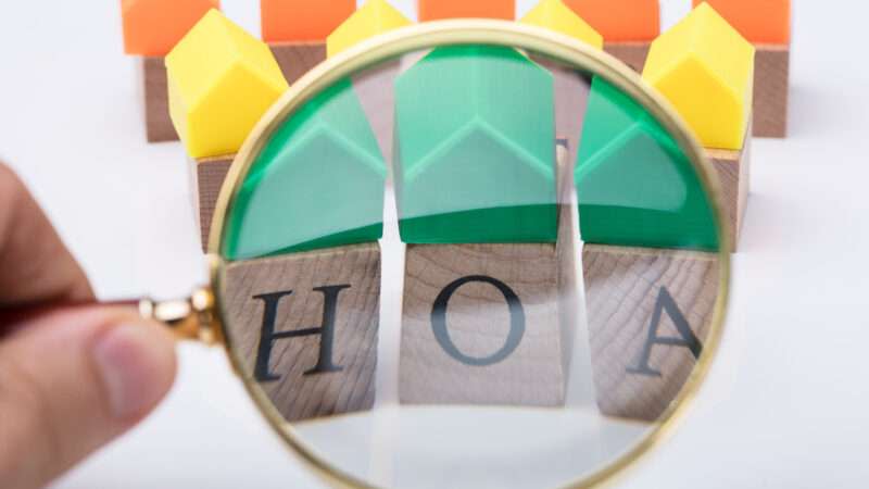 A magnifying glass in front of wooden letters spelling HOA and a row of multicolored houses behind it against a white background