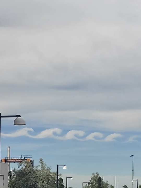 Sky with distinctive wave-like clouds above a cityscape, featuring streetlights and industrial buildings. No persons are in the image