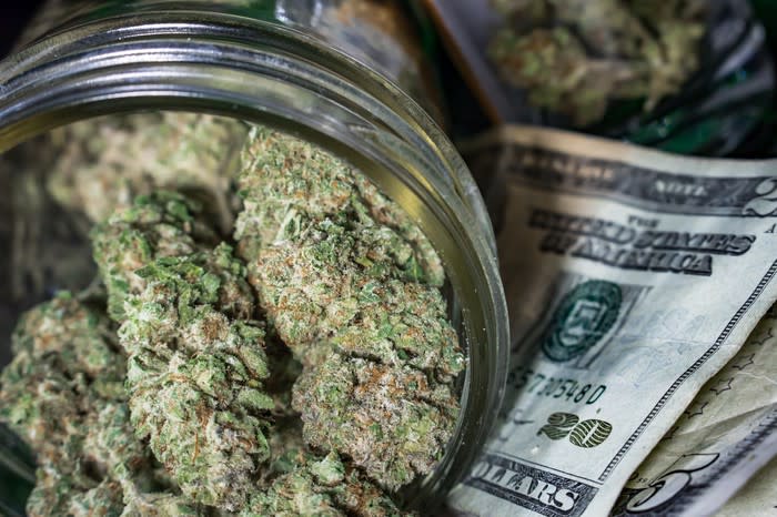 A clear jar packed with dried cannabis buds that's lying atop a small pile of cash.