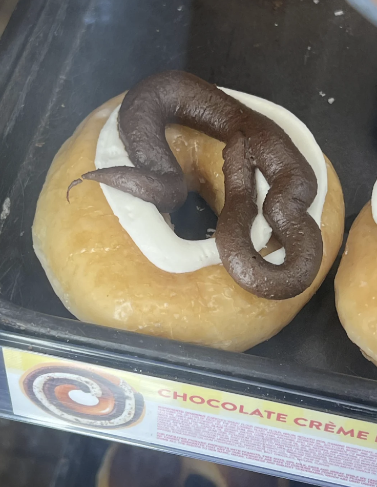 A doughnut with white icing is topped with what appears to be chocolate frosting, shaped like feces. The label reads "CHOCOLATE CRÈME."