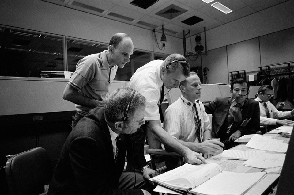 black and white photo of six men sitting or standing near a table with lots of computer printouts and notebooks on it.