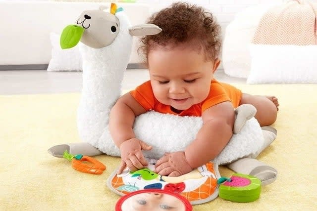 Infant sitting on a plush tummy wedge toy with various textures and tags, suitable for age 0+