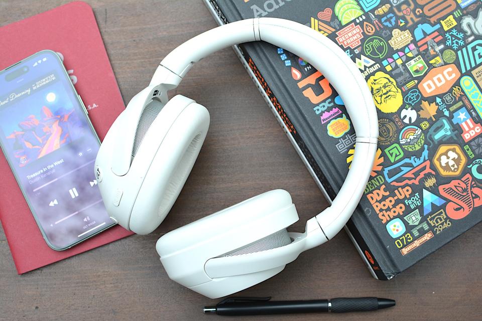 <p>White headphones sitting on a wooden table from above with a red notebook and phone on the left and a black book with logos on the cover on the right/underneath.</p>
