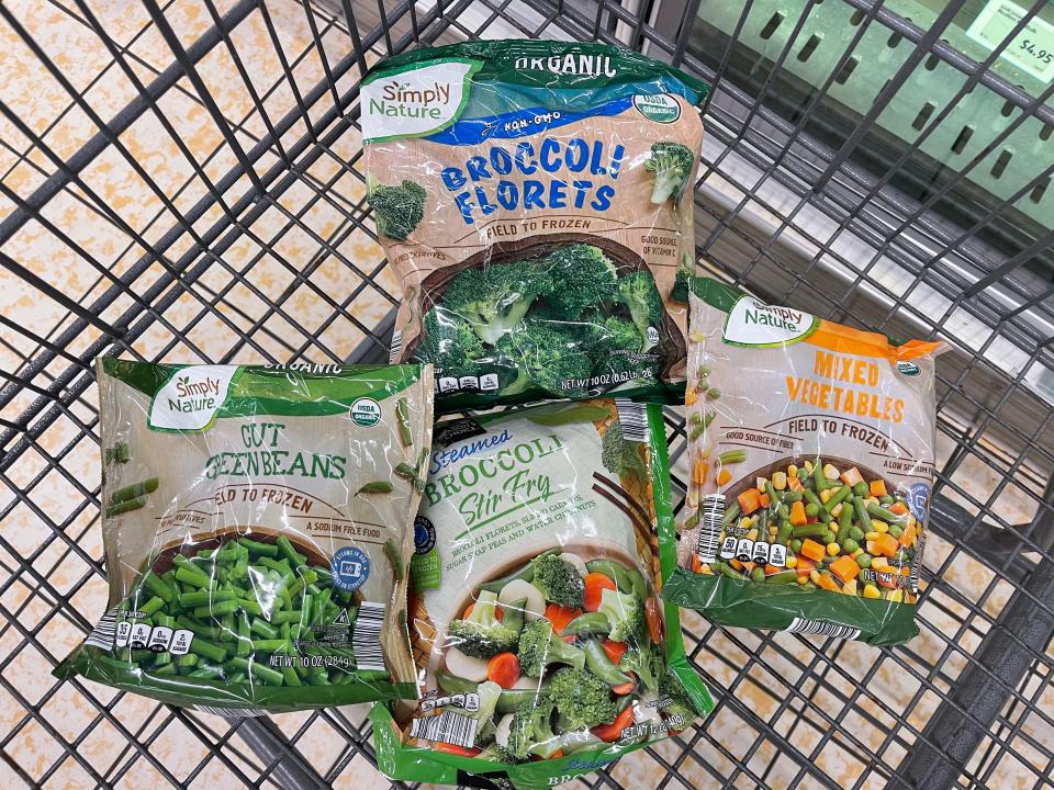 Four bags of Simply Nature frozen vegetables in a shopping cart: cut green beans, broccoli florets, steamed-broccoli stir fry, and mixed vegetables.