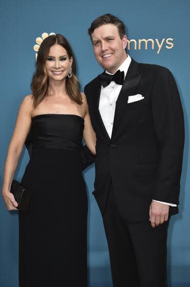 A woman with wavy hair and a black strapless dress smiling and posing next to a man with brown hair in a black tuxedo