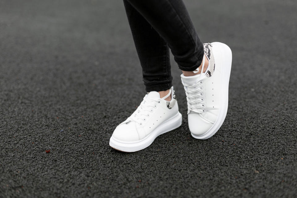 Female legs in stylish black jeans in white leather sneakers with snake pattern on the asphalt in the city. Young women on a walk. Fashionable women's shoes. New collection. Close-up.