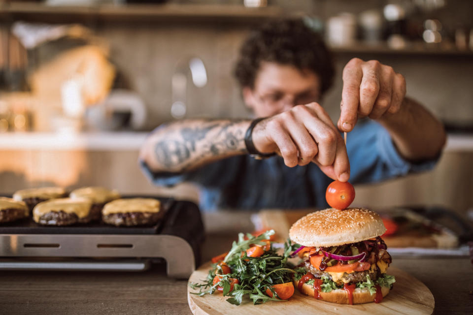 Young Man Decorating Delicious Hamburgers In Kitchen