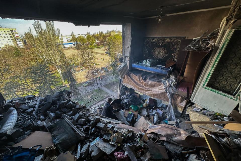 A view of a tree-lined park is seen through the missing wall of a rubble-filled apartment.