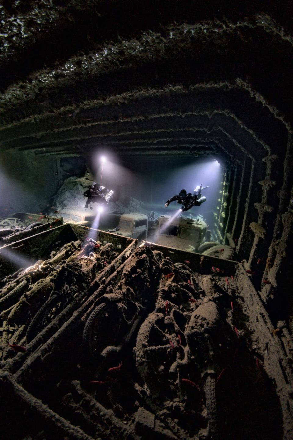 Two divers explore the interior of the wreck of the SS Thistlegorm in the Egyptian Red Sea, their torches illuminating rows of silt-covered motorcycles inside the ship. The image is a winner in the 2024 Underwater Photographer of the Year Awards.