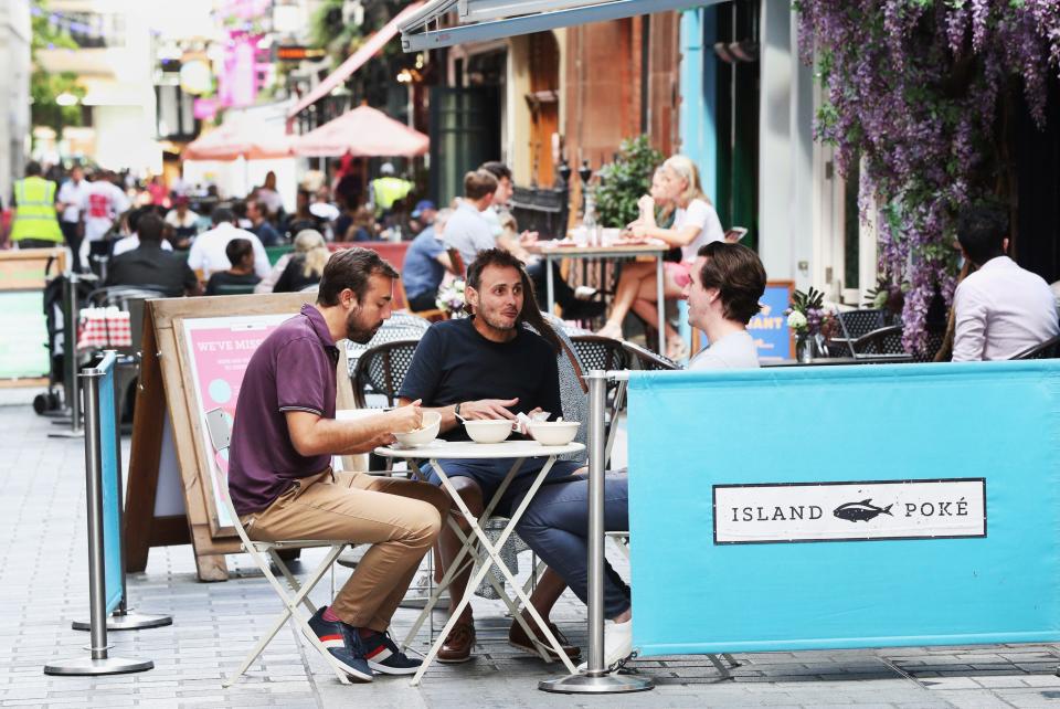 Three people sit at an outside table at Island Poke, a poke restaurant in London, with a light blue banner with "Island Poke" on it sit in the foreground.