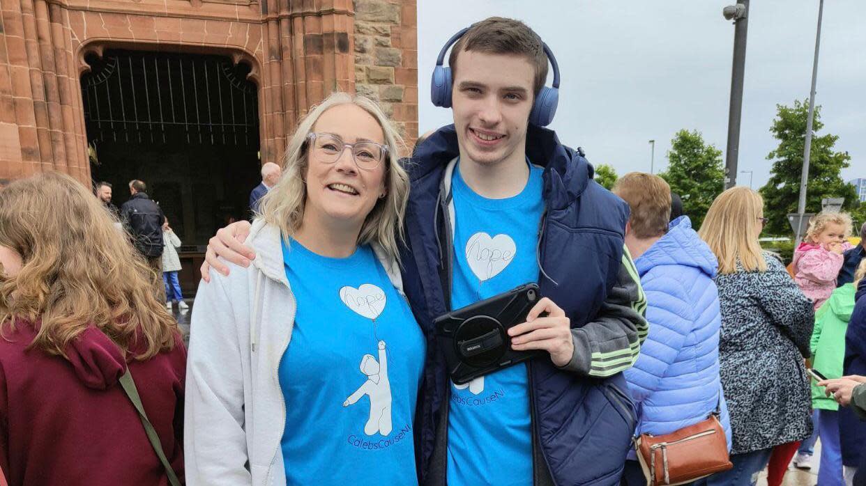 A smiling Caroline Campbell is embraced by her teenage son Oscar as they both stand outside the Guildhall in Derry. They are both wearing blue t-shirts, Caroline is wearing a grey hoodie while Oscar, who is also smiling, is wearing a navy jacket and headphones on his head