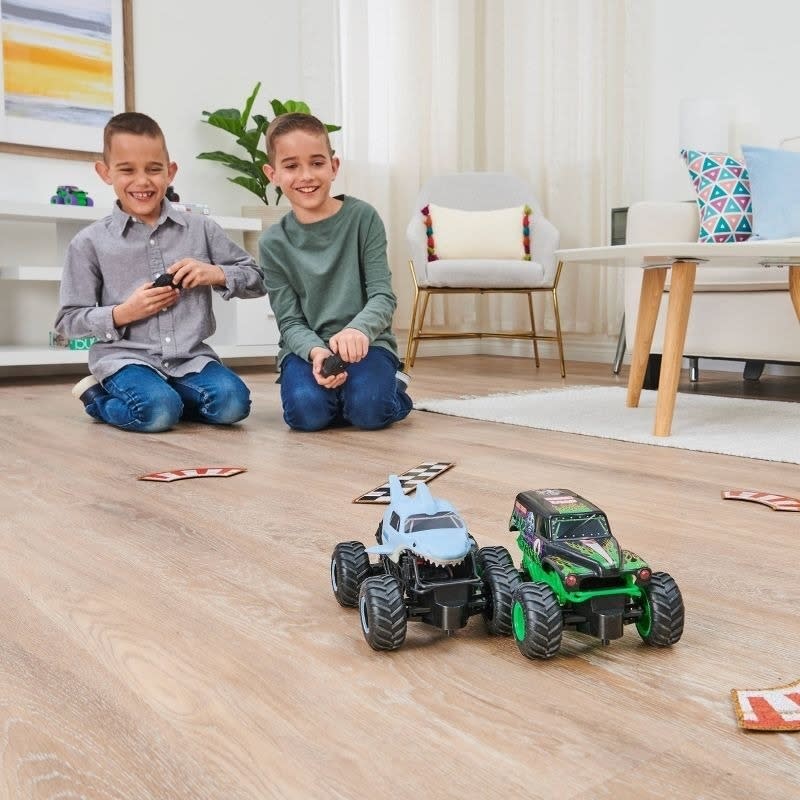 Two children playing with remote-controlled monster trucks on a hardwood floor