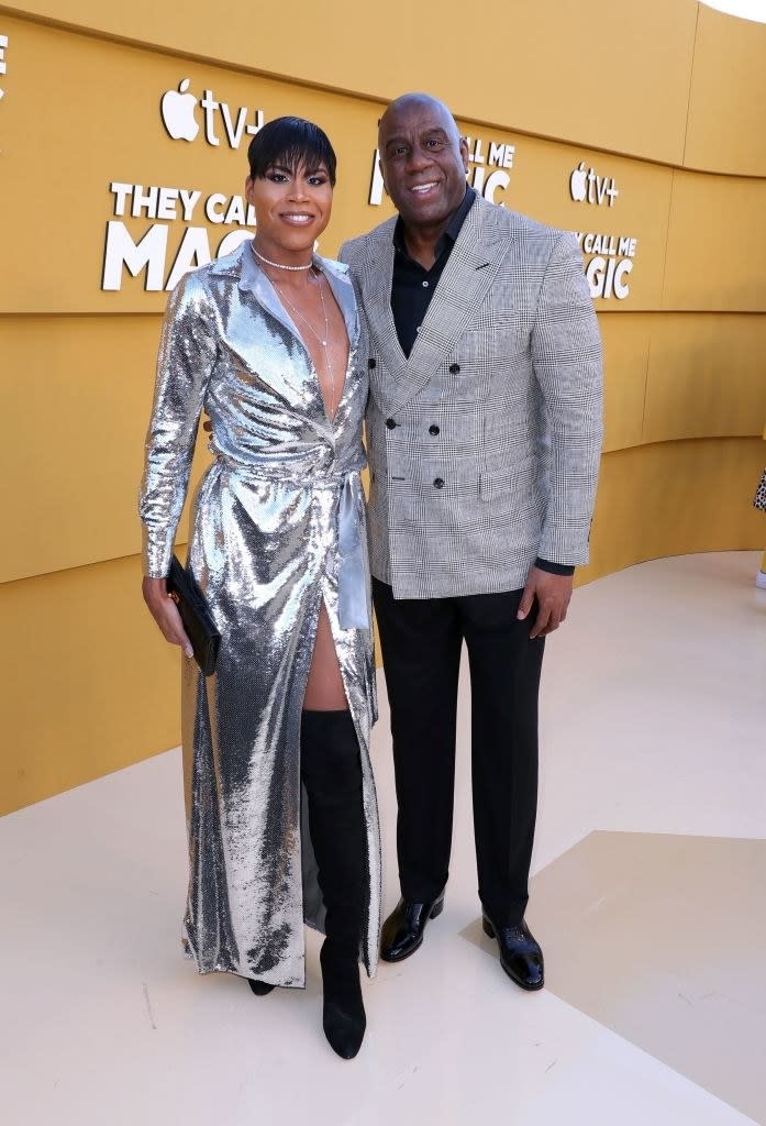 EJ in a silvery gown with front slit and thigh-high boots standing with a smiling Magic, who is wearing a suit and has his arm around EJ