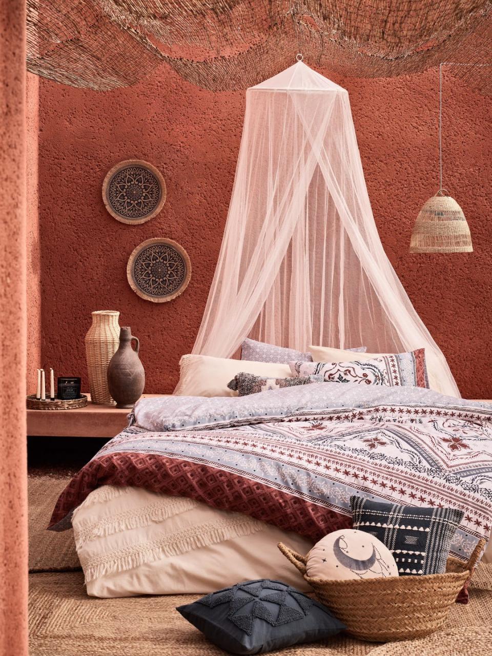 layered bed with canopy and piled with cushions against a rough terracotta wall