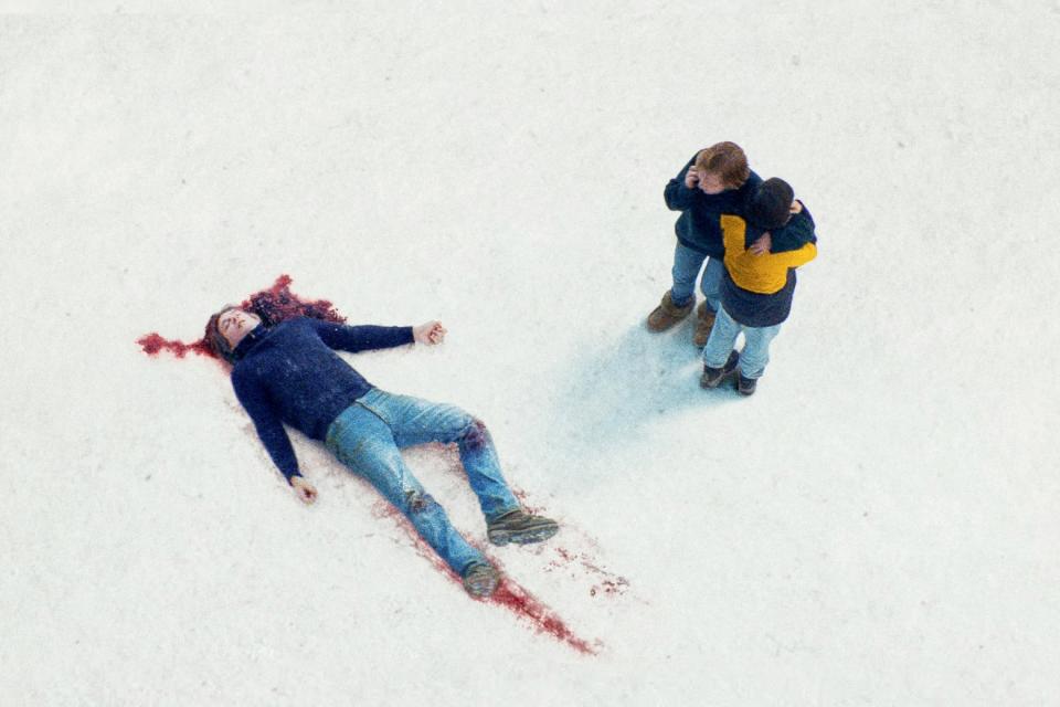 A man lies in the snow with lots of blood, two people stand over him. 
