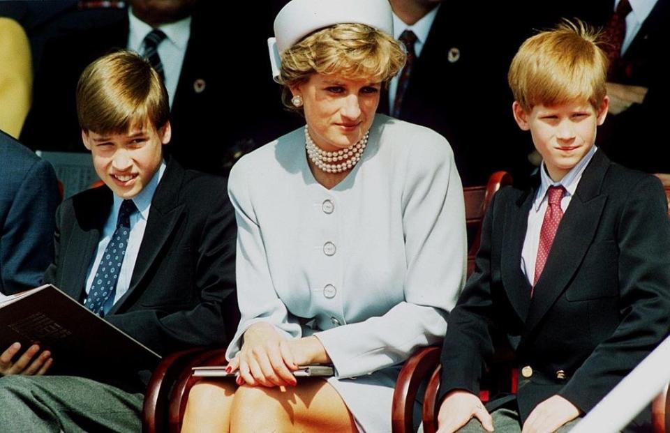 From left to right: A young Prince William, Princess Diana, and a young Prince Harry sitting in a row at an outdoor event