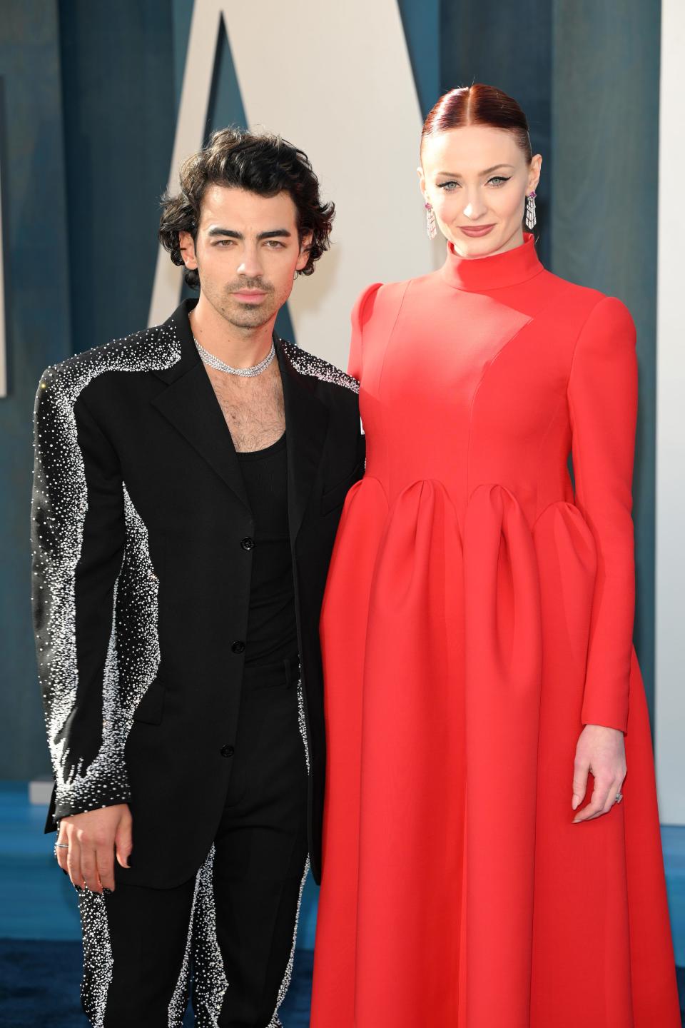 Joe in a black blazer, tank top, and pants. The silhouette of the jacket and pants are outlined in silver clustered crystals. He's wearing a matching crystal necklace. Sophie is wearing a turtleneck long sleeve red dress that poofs out over her stomach, enhancing her baby bump.