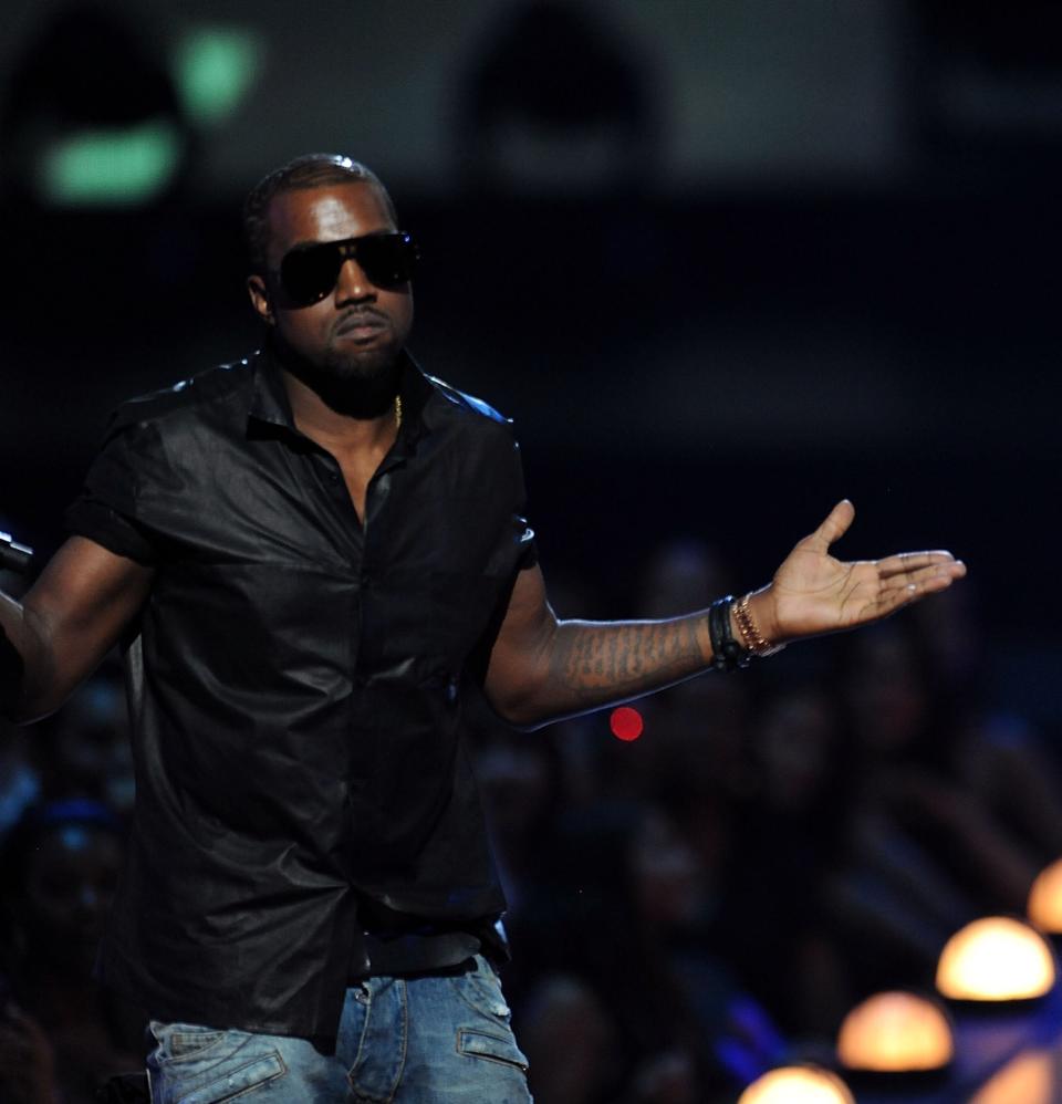 Kanye West, in sunglasses and black shirt, gestures with arms outstretched on stage while Taylor Swift, in a sparkling dress, holds a trophy and looks on