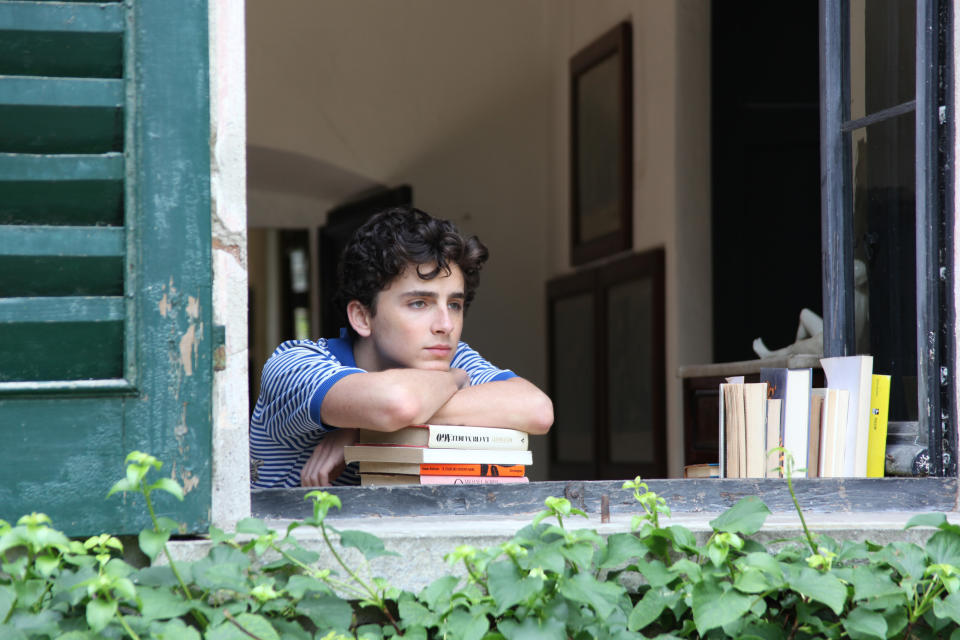 Timothée Chalamet leans on a stack of books while looking out of an open window in a contemplative pose