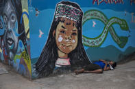 A homeless man sleeps against a wall adorned with a mural featuring a Shipibo Indigenous girl and Amazon rainforest animals, in Pucallpa, in Peru's Ucayali region, Wednesday, Sept. 2, 2020. Peru is home to one of Latin America's largest Indigenous populations, whose ancestors lived in the Andean country before the arrival of Spanish colonists. (AP Photo/Rodrigo Abd)