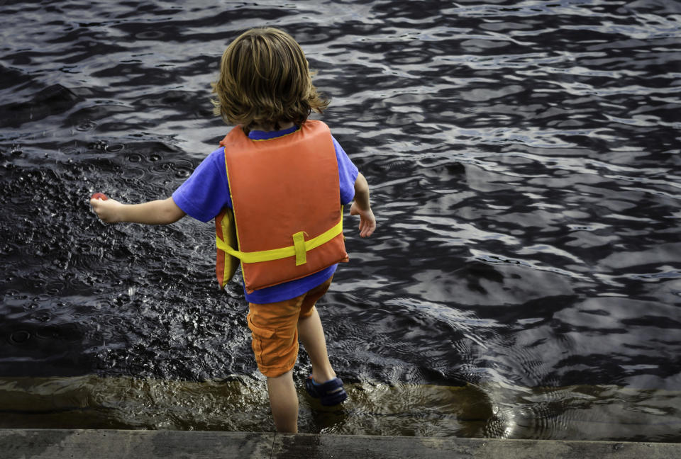 A child wearing a life jacket stands at the edge of the water, holding a toy while wading