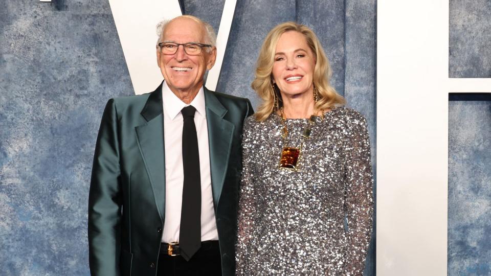 jimmy buffett and his wife jane hold hands while smiling for photos, he wears a teal shiny suit jacket with a white collared shirt, black tie and black pants, she wears a silver glittering long sleeve gown with a large amber colored necklace