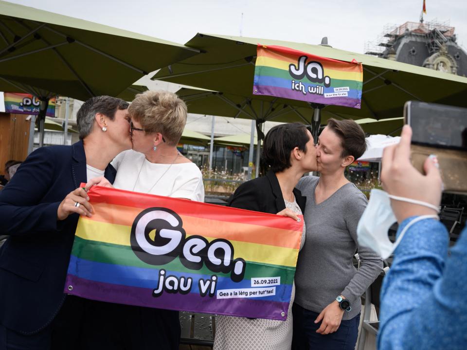 Two same-sex couples kiss while holding a pride flag.