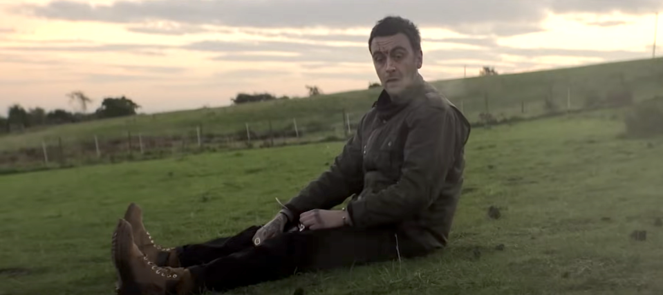 joseph gilgun in brassic series 5, a man sits in the middle of a field with cuts on his face