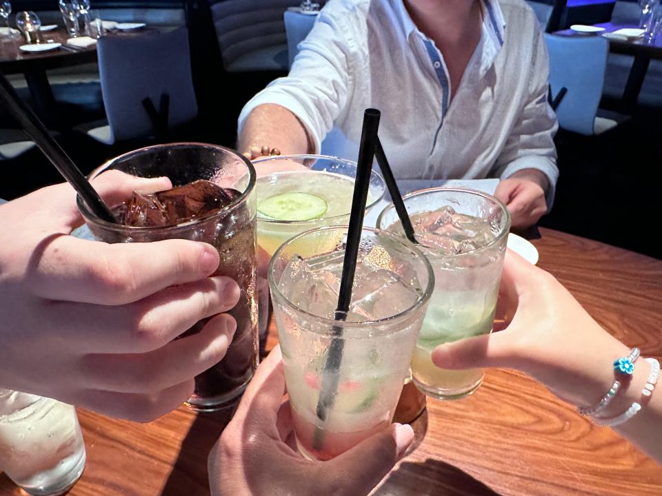 Four drinks clinking together at a wooden table with a man in a light-blue button-down across the table