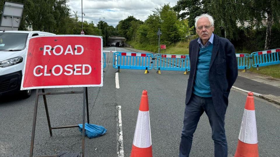 Councillor Richard Kemp wears a navy blue jacket and trousers with lighter blue shirt and jumper, standing next to the cordoned off road, with traffic cones and a 'road closed' sign next to him