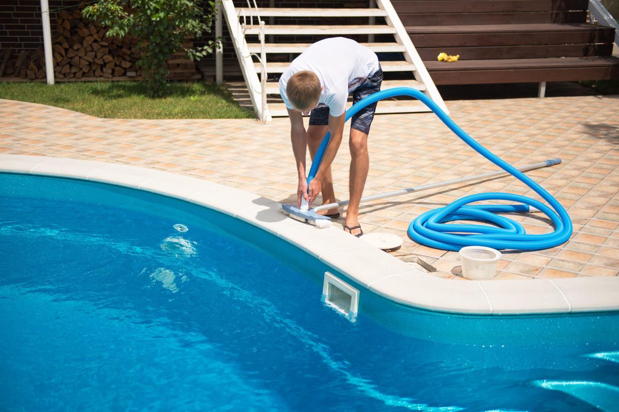 Man cleaning an in-ground swimming pool with a pool vacuum cleaner, standing while about to put in the vacuum cleaner with brick surrounding the pool, steps and lawn in the background