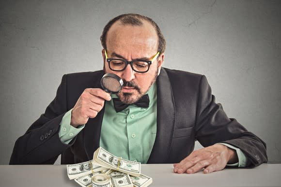Bespectacled businessman looking at pile of money through magnifying glass