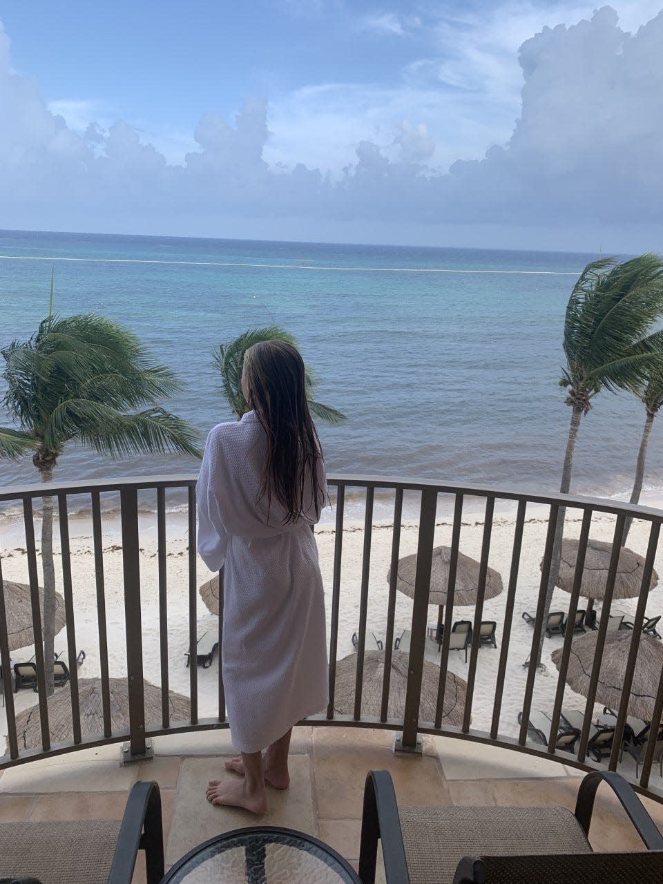 A girl standing on a balcony with her back to the camera, facing the beach, wearing a robe.