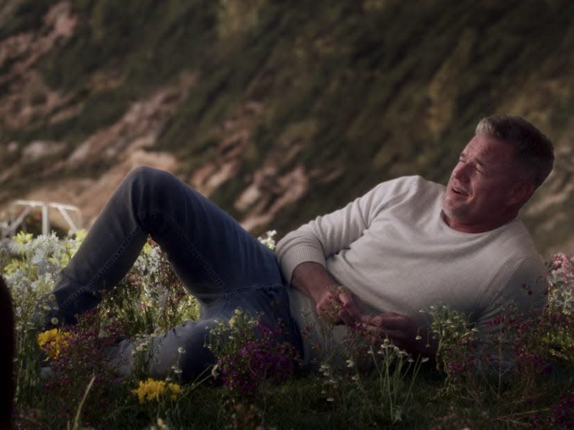 Mark sitting in the flowers on a beach wearing a white sweater and jeans on Greys Anatomy