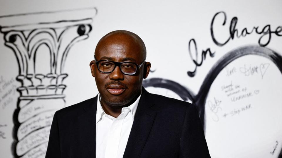British Vogue editor-in-chief Edward Enninful, wearing black rimmed glasses, a white shirt and dark blazer. He's looking at the camera. The background is white with black writing and artwork on the wall.