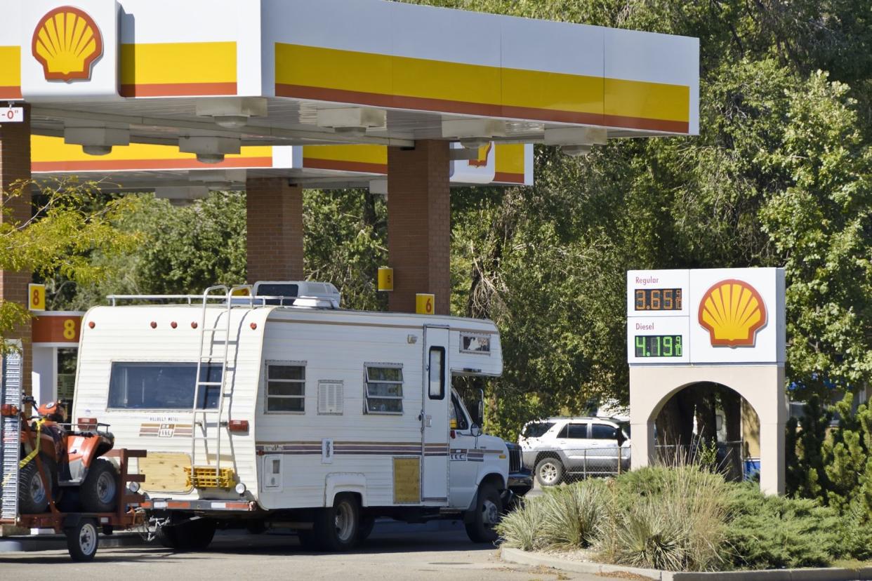 An RV at a pump at a Shell gas station, Fort Collins, Colorado, trees surround the side of it, on a sunny day