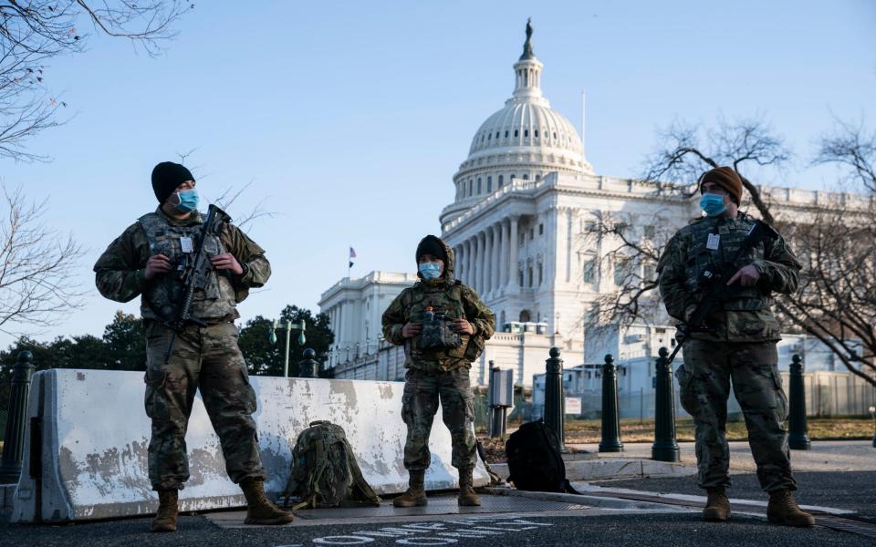 Three members of the US National Guard stand ready in front of a chipped concrete portable roadblock in the cold morning light, as the vast neoclassical bulk and ivory-hued dome of the US Capitol building looms behind them, half bright and half in blue shadow. All three soldiers wear pale blue surgical masks, woollen beanie caps and military fatigues with bulletproof vests and numerous pockets. Two are carrying assault rifles, probably M4 carbines, but showing proper trigger discipline. - Sarah Silbiger/Getty