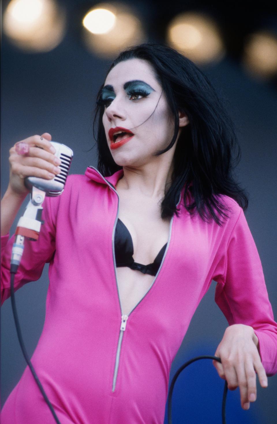 PJ Harvey, with thick brows and dark hair, on stage in bold makeup, singing into a vintage microphone, wearing a vibrant jumpsuit