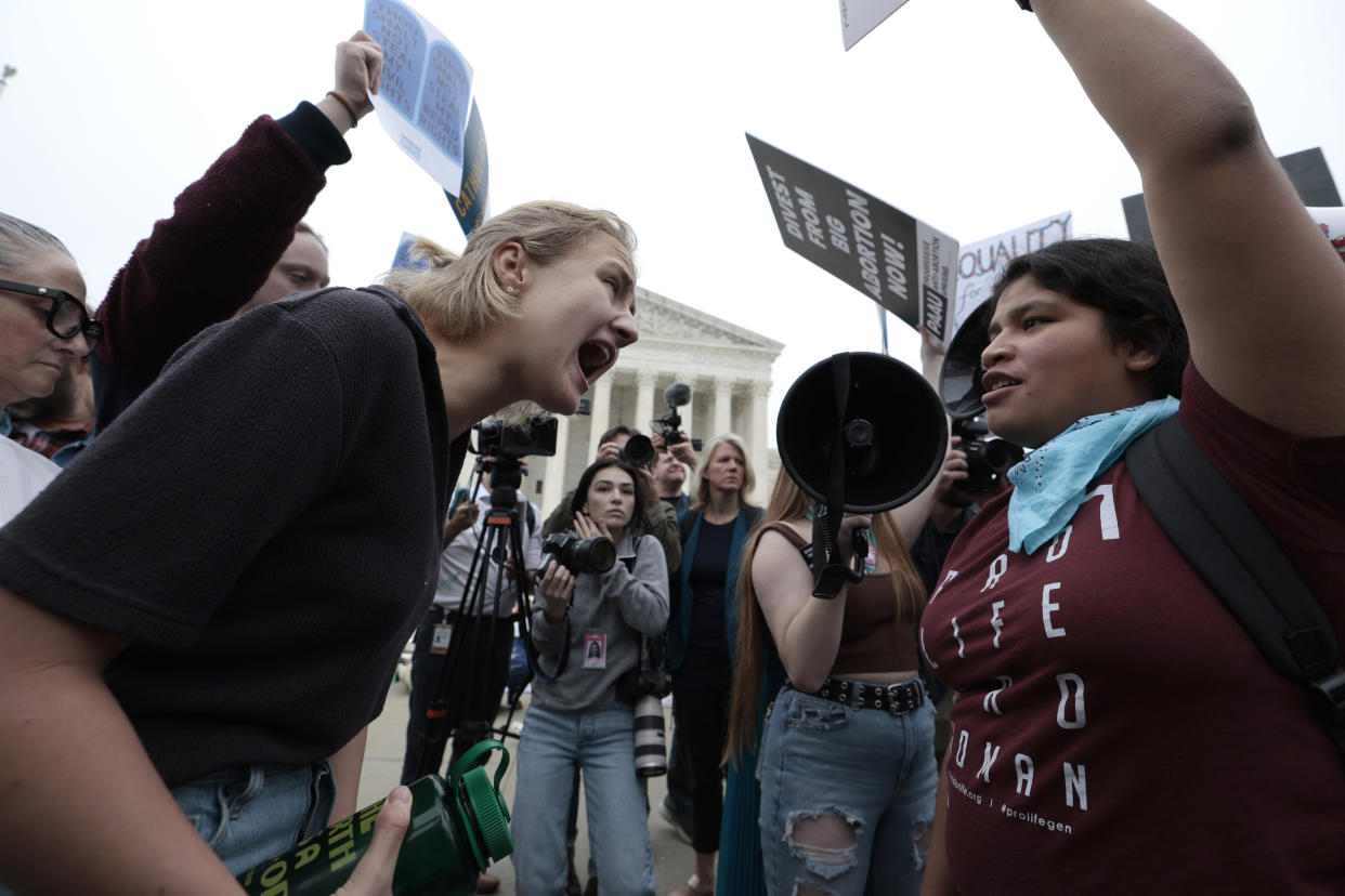 A blonde young woman shouts at another young woman, as someone holds a bullhorn behind them. Photographers are capturing the scene, with the Supreme Court in the background.n.