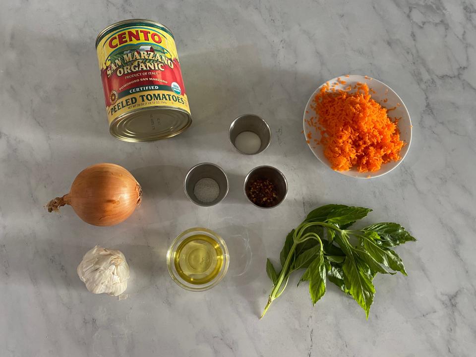 Can of peeled tomatoes, basil, garlic, onion, oil, bowl of grated carrots, and spices on a counter top